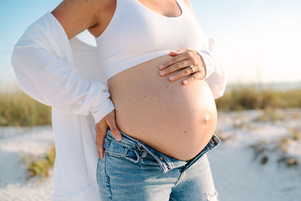 Maternity photo on the beach, woman wearing jeans for maternity photoshoot holding baby bump